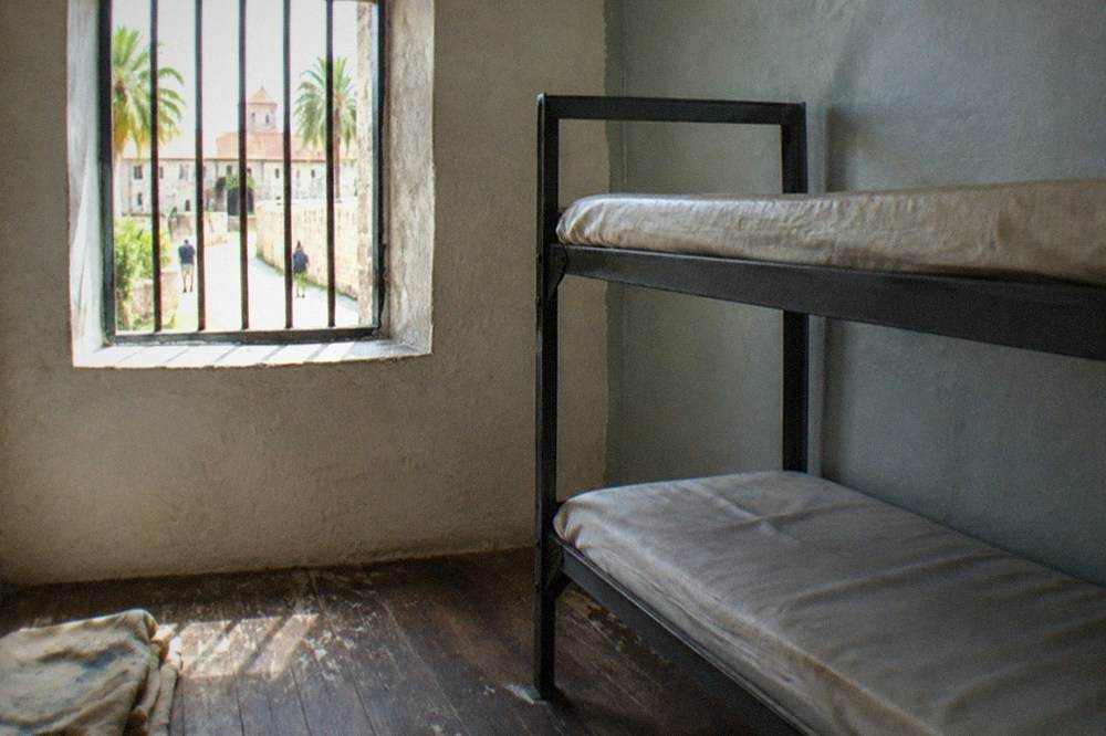 A historic jail cell with a narrow iron bunk and worn wooden floor