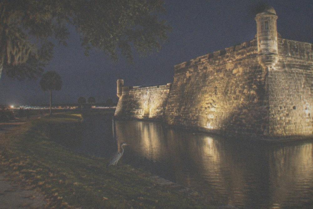 A dark historic fortress wall lit by floodlights across a water moat at night