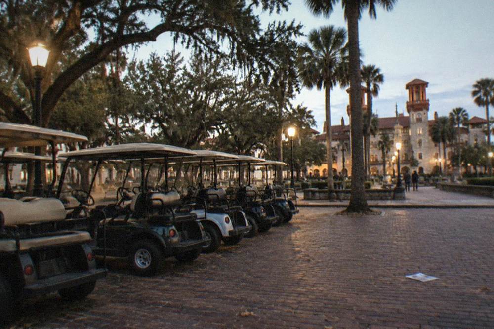 A row of parked golf carts under streetlights near a plaza bordered by old stone buildings