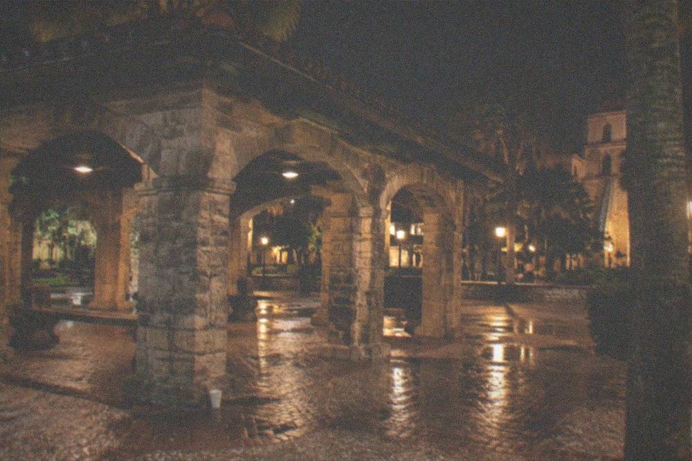A plaza at night with an old stone market pavilion lit by streetlamps and palm trees