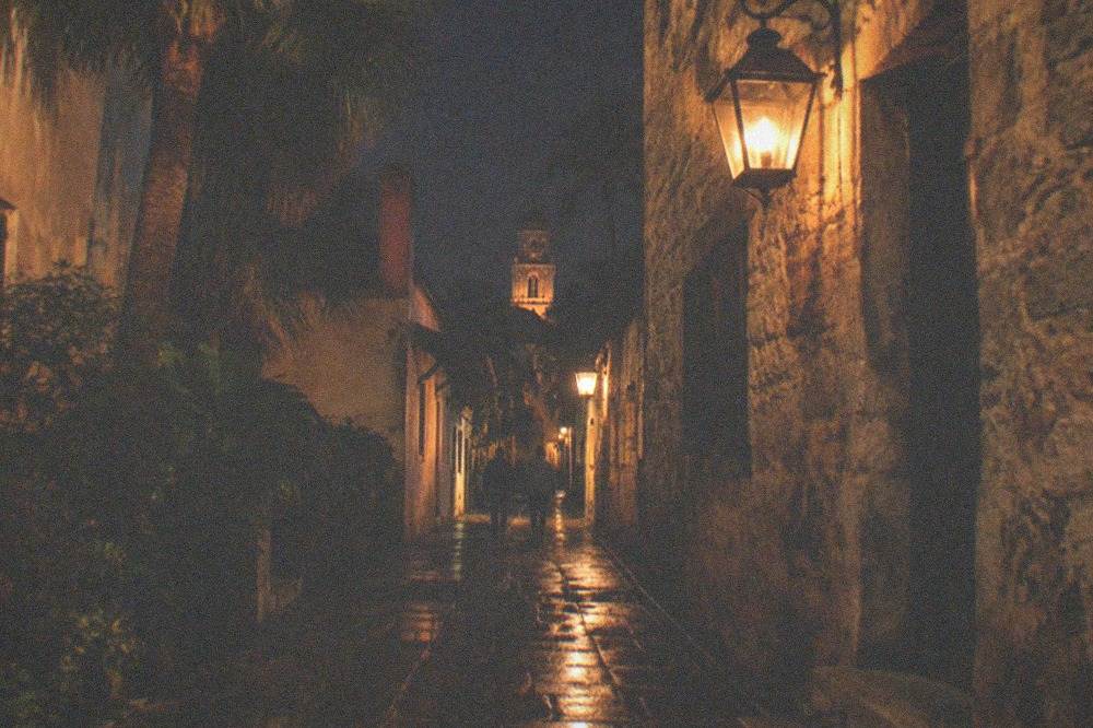 A narrow historic lane at night with coquina walls, hanging lanterns and deep shadows between buildings